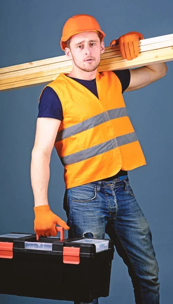 ヘルメット、ヘルメットの男は、ツールボックスと木製の梁、灰色の背景を保持します。大工、木工家、労働者、穏やかな顔の肩に木の梁を運ぶのビルダー。木工のコンセプト — ストック写真