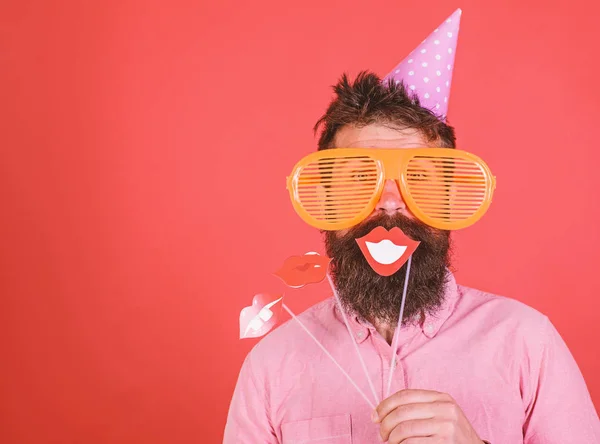 El tipo con sombrero de fiesta lo celebra, posando con accesorios fotográficos. Hipster en gafas de sol gigantes celebrando. Hombre con barba en la cara alegre sostiene los labios sonrientes en palos, fondo rojo. Concepto de diversidad emocional —  Fotos de Stock