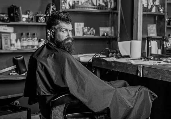 Человек с бородой в черном плаще сидит в парикмахерском кресле перед зеркалом. Человек с бородой клиент хипстерской парикмахерской. Концепция парикмахерской. Хипстер с бородой ждет парикмахера и стрижку — стоковое фото