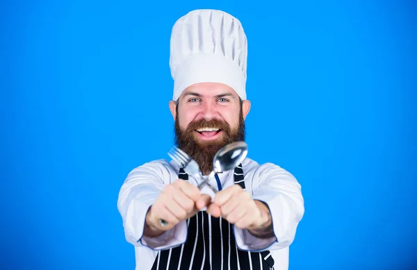 Homem bonito com barba segura utensílios de cozinha no fundo azul. Conceito de processo de cozinha. Vamos experimentar o prato. Chef faminto pronto para experimentar comida. Está na hora de provar. Chef feliz sorridente rosto segurar colher e garfo — Fotografia de Stock