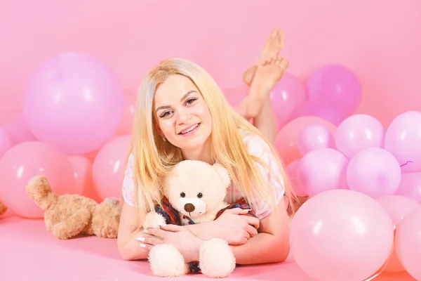 Girl in pajama, domestic clothes lay near air balloons, pink background. Blonde on smiling face relaxing with teddy bear toy. Birthday girl concept. Woman cute celebrate birthday with balloons.