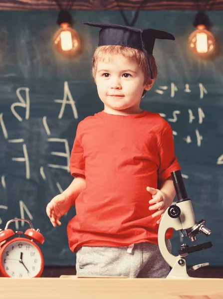 Concepto de graduación de jardín de infantes. Primero interesado en estudiar, educación. Niño, pupila sonriente cerca del microscopio. Niño con gorra de graduado listo para ir a la escuela, pizarra en backgro — Foto de Stock