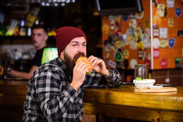 Enjoy taste of fresh burger. Hipster hungry man eat burger. Man with beard eat burger menu. Brutal hipster bearded man sit at bar counter. Cheat meal. High calorie food. Delicious burger concept