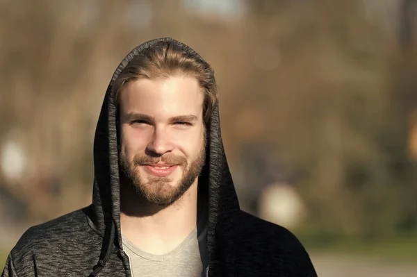 Mann mit Bart lächelt in Kapuze an einem sonnigen Tag. Bärtiger Mann trägt lässiges Sweatshirt im Freien. Mode-Macho lächelt in stylischer Sportbekleidung. Lebensstil für aktive und gesunde Menschen, Sport — Stockfoto