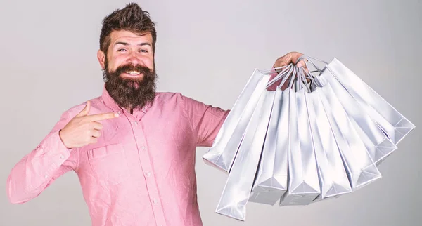 Concepto de recomendación. Chico de compras y apuntando a las bolsas. El hombre con barba y bigote lleva un montón de bolsas de compras, fondo gris. Hipster en la cara feliz recomienda comprar —  Fotos de Stock