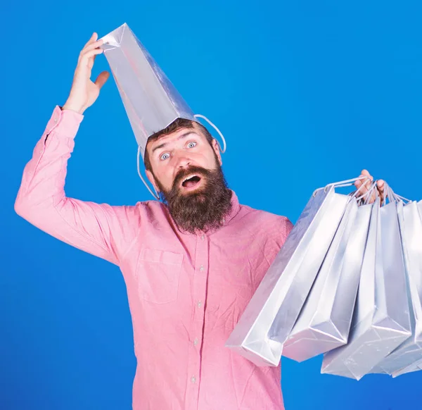 Hombre con barba y bigote lleva bolsas de compras, fondo azul. Concepto de compras. Hombre de compras en temporada de ventas con descuentos. Hipster en la cara sorprendida con bolsa en la cabeza es adicto a las compras —  Fotos de Stock