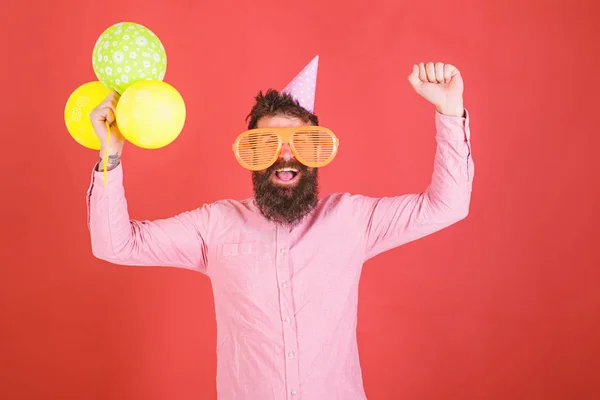 Hipster in gigantische zonnebril viert verjaardag. Man met baard en snor op blij gezicht houdt stelletje lucht ballonnen, rode achtergrond. Guy in feest hoed viert vakantie. Viering concept — Stockfoto