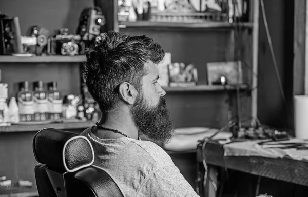 Homem com barba e bigode senta-se na cadeira de cabeleireiro, suprimentos de beleza no fundo. Hipster com barba espera por barbeiro e corte de cabelo. Homem com barba cliente da barbearia hipster. Conceito de barbearia — Fotografia de Stock