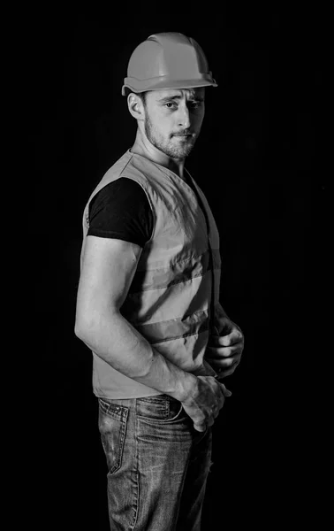 Attractive builder concept. Builder in helmet posing confidently. Man in helmet, hard hat wears orange vest, black background. Worker, contractor, builder on serious face with muscular biceps — Stock Photo, Image