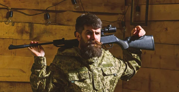 Masculinity concept. Hunter, brutal hipster on strict face with gun ready for hunting. Man, gamekeeper with beard wears camouflage clothing, carries rifle on shoulders, wooden interior background