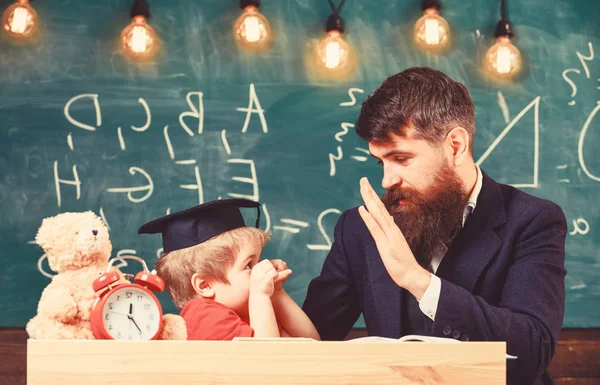 Kid vrolijke afleiden tijdens het studeren, aandacht tekort. Leraar en leerling in mortarboard, schoolbord op achtergrond. Speels kind concept. Vader met baard, leraar leert zoon, kleine jongen — Stockfoto