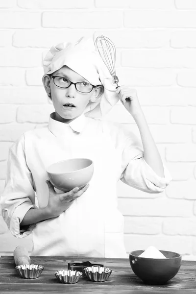 Jeune garçon chef cuisinier mignon en uniforme blanc et chapeau sur le visage surpris avec des lunettes debout près de la table avec des moules à rouleau pour gâteaux et tenant bol turquoise sur fond de mur de brique — Photo