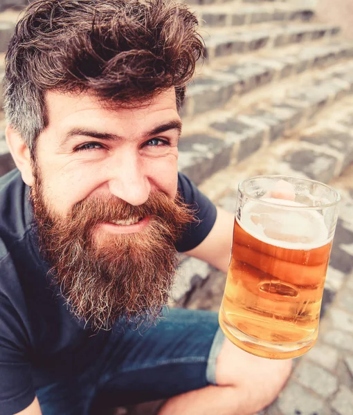 Un tipo descansando con cerveza de barril. Hipster en la cara sonriente alegre bebe cerveza al aire libre. Concepto de celebración. Hombre con barba y bigote sostiene vidrio con cerveza mientras se sienta en escaleras de piedra, desenfocado — Foto de Stock