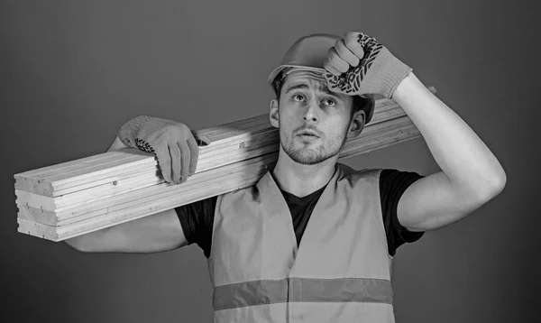 Man in beschermende handschoenen houdt vizier van beschermende harde hoed, grijze achtergrond. Timmerman, schrijnwerker, sterke bouwer op ernstige gezicht draagt houten balk op de schouder. Veiligheid en bescherming concept — Stockfoto