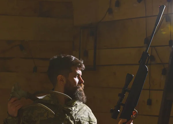 Hunter concept. Macho on strict face at gamekeepers house ready for hunting. Hunter brutal with gun and horns of deer, lighted in darkness. Man with beard wears camouflage clothing, dark background — Stock Photo, Image