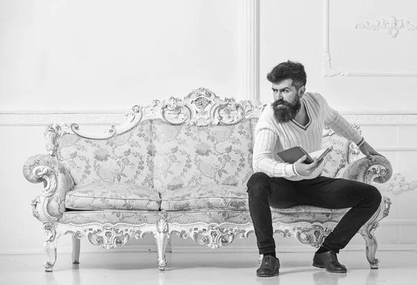 Man met baard en snor zit op barokstijl sofa, wachtruimten boek, witte muur achtergrond. Macho op strikte gezicht klaar met het boek te lezen. Beschouwingen over literatuur concept. Guy denken over literatuur — Stockfoto