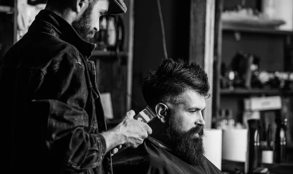 Hipster client getting haircut. Barber with hair clipper works on haircut of bearded guy, retro barbershop background. Barber with clipper trimming hair on nape of client. Hipster hairstyle concept