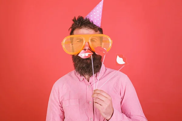 O tipo de chapéu de festa festeja, posando com adereços fotográficos. Conceito de diversidade emocional. Homem com barba na cara alegre mantém lábios sorridentes em paus, fundo vermelho. Hipster em óculos de sol gigantes comemorando — Fotografia de Stock