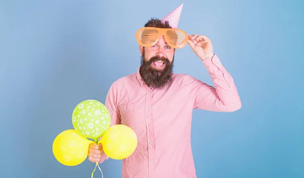 Homem barbudo alegre com grande sorriso usando óculos enormes e boné de aniversário. Hipster feliz com barba aparada e bigode em camisa rosa posando com balões amarelos e verdes em fundo azul — Fotografia de Stock