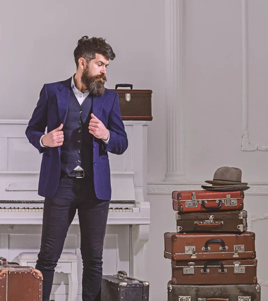 Macho con estilo en la cara reflexiva de pie cerca de pila de maleta vintage. Hombre, viajero con barba y bigote con equipaje, lujoso fondo interior blanco. Equipaje y concepto de viaje — Foto de Stock