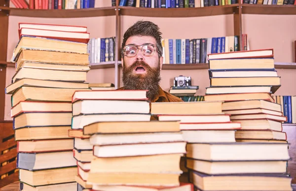 Man, nerd on surprised face between piles of books in library, bookshelves on background. Nerd concept. Teacher or student with beard wears eyeglasses, sits at table with books, defocused