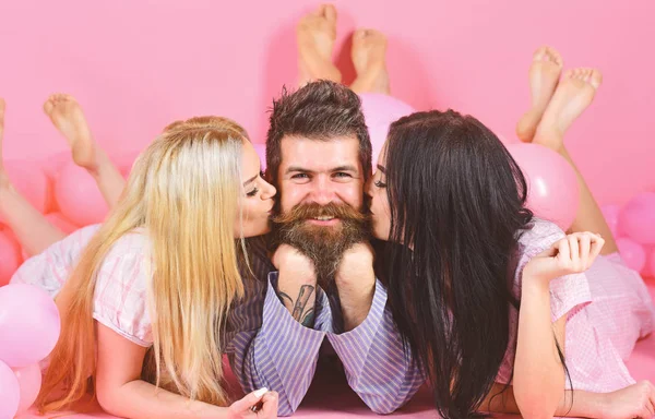 Alpha male concept. Threesome lay near balloons, happy guy on smiling face. Man with beard and mustache attracts blonde and brunette girls. Girls fall in love with macho, kissing, pink background