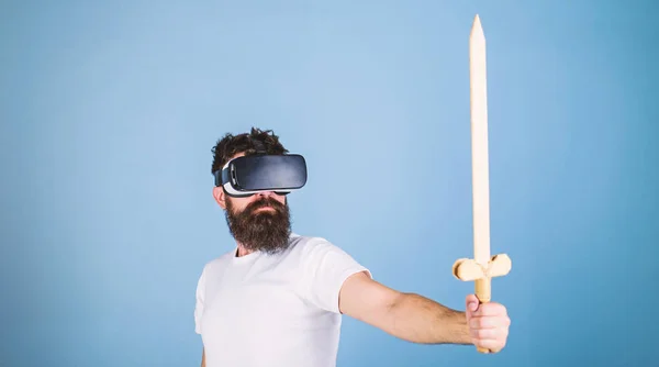 Gamer concept. Guy with head mounted display holds sword, play fighting game in VR. Hipster on serious face enjoy play game in virtual reality. Man with beard in VR glasses, light blue background