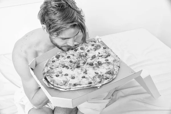 Man likes pizza for breakfast. Bachelors nutrition. Man bearded handsome guy eating cheesy food for breakfast in bed. Guy holds pizza box sit bed in bedroom or hotel room. Food delivery service