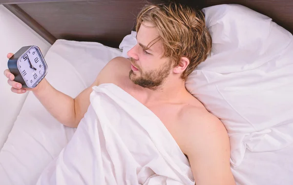 Homem sonolento sonolento sem barba rosto coberto com cobertor tendo descanso. Mantenha o horário de dormir na mesma hora de dormir e acorde. Conceito de hábitos de regime de sono. Homem unshaven lay desperta cama segurar despertador — Fotografia de Stock