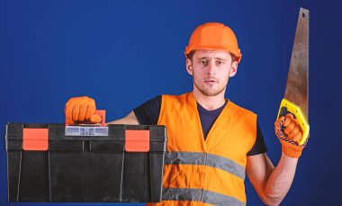 Marangoz kavramı. Kask, kask içinde adam araç kutusu taşır ve el testeresi, mavi arka plan tutar. İşçi, repairer, tamirci üzerinde ciddi bir yüz taşıyan araç, onarım için kopya alanı hazır