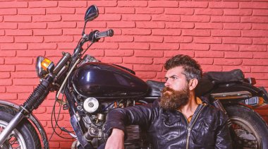 Sakallı, motorcu motorlu bisiklet garaj, tuğla duvar arka plan yakınındaki deri ceketli adam. Yenilikçi, vahşi motorcu deri ceketli dalgın yüzünde oturuyor, motosiklet üzerinde eğilir. Bisikletçileri yaşam tarzı kavramı