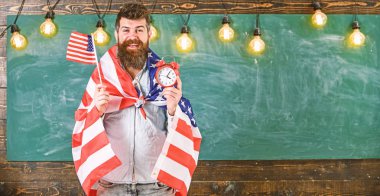 Amerikalı öğretmen Amerikan bayrakları ile çalar saat tutar. Amerikan eğitim sistemi kavramı. Mutlu yüz sakallı adam bayrak ABD ve saat sınıf, arka plan, kopya spac kara tahta tutar