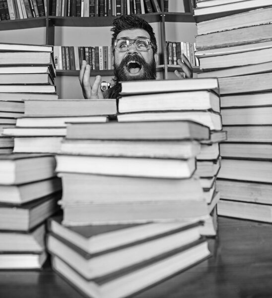 Teacher or student with beard wears eyeglasses, sits at table with books, defocused. Scientific discovery concept. Man on excited face between piles of books in library, bookshelves on background