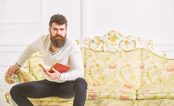 Férfi szakáll és bajusz ül a barokk stílusú kanapé, tartja a könyvet, fehér háttér. Gondolatok az irodalom fogalma. A fickó gondolkodó irodalom. Macho-a szigorú arcát befejezte az olvasást a könyv — Stock Fotó