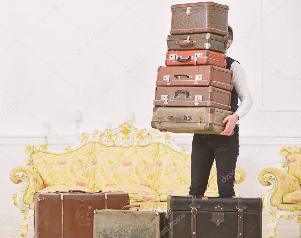 Butler and service concept. Macho, elegant porter carries heavy pile of vintage suitcases. Man with beard and mustache wearing classic suit delivers luggage, luxury white interior background