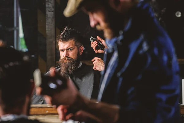 Barber with hair clipper works on hairstyle for man with beard, barbershop background. Haircut concept. Hipster client getting haircut. Barber styling hair of brutal bearded client with clipper