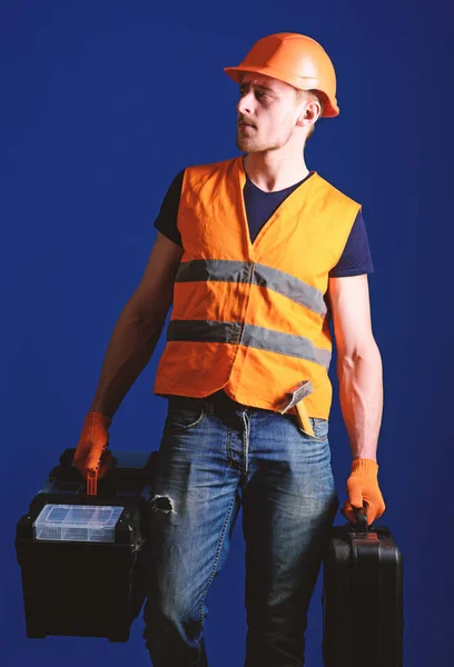 Чоловік в шоломі, жорсткий капелюх тримає коробку інструментів і валізу з інструментами, синій фон. Працівник, майстер, ремонтник, будівельник на спокійному обличчі несе сумки з професійними інструментами. Концепція професійного ремонтанта — стокове фото