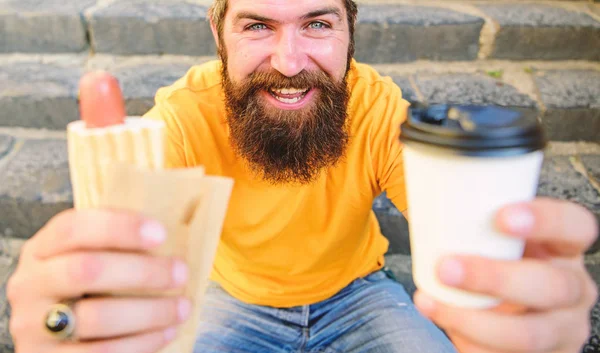 Fast food para almoço ideia comum. Hipster desfrutar de cachorro-quente e beber bebida copo de papel. Homem barbudo desfrutar rua comida escadas fundo. Hipster come cachorro-quente e bebe café. Conceito de hora de almoço — Fotografia de Stock