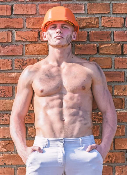 Builder with muscular torso and helmet, brick wall and sky on background.  Athlete with nude torso in hard hat, copy space. Sexy builder concept.  Builder with strong muscular body covered with dust