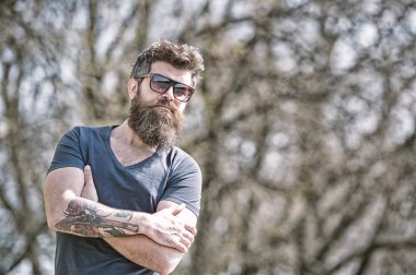 Ciddi sakallı adam kollarını ile ayakta geçti. Yakışıklı adam açık havada poz trendy güneş gözlüğü takıyor dövme ile