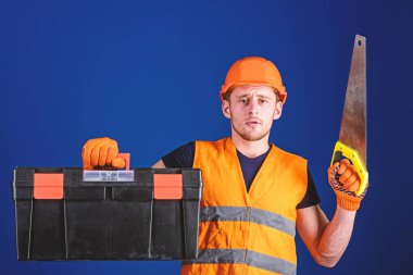 Marangoz kavramı. İşçi, repairer, tamirci üzerinde ciddi bir yüz taşıyan araç, onarım, kopya alanı için hazır. Kask, kask içinde adam araç kutusu taşır ve el testeresi, mavi arka plan tutar