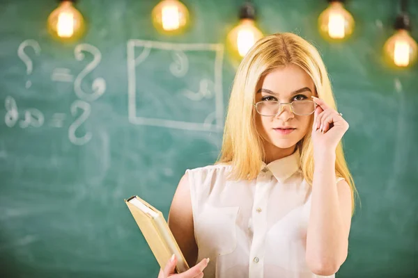 学生、教師に見える眼鏡に自信を持って、デフォーカスの背景に黒板の教室で立っています。魅力的な講師のコンセプトです。本を持つ女性のレッスンの準備、観客の見つめる — ストック写真