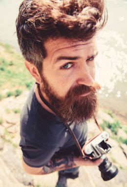 Sakal ve bıyık tutar kamera, güneşli, su yüzeyine arka plan açık olan adam. Fotoğrafçı kavramı. Eski moda kamera hipster katı yüz tutar. Nehir kenarında doğa çekim adam
