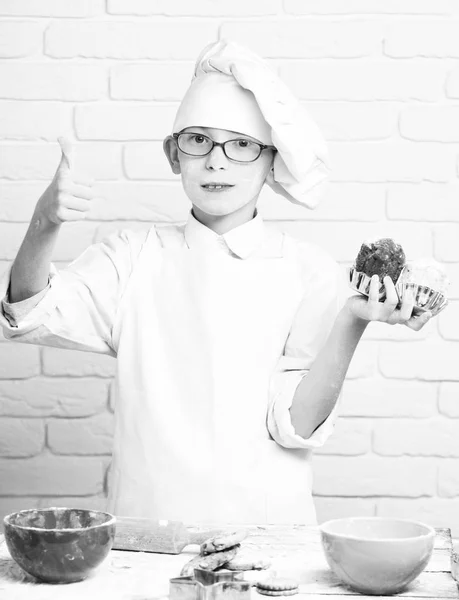 Young boy cute cook chef in white uniform and hat on stained face flour with glasses standing near table with colorful bowls and holding chocolate cakes on brick wall background — Stock Photo, Image