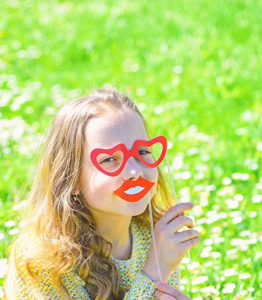 Love from first sight concept. Child posing with cardboard heart shaped eyeglasses and smiling mouth. Girl on happy face spend leisure outdoors. Girl sits on grass at grassplot, green background