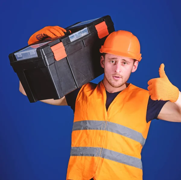 Trabalhador, reparador, reparador, construtor no rosto confiante carrega caixa de ferramentas no ombro. Homem de capacete, chapéu duro segura caixa de ferramentas e mostra gesto polegar para cima, fundo azul. Conceito de consulta de reparação — Fotografia de Stock