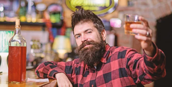 Guy spend leisure in bar, defocused background. Hipster with beard holds alcoholic beverage. Cheers concept. Man drinks whiskey or cognac. Man on relaxed face sits near bar counter, raising up glass