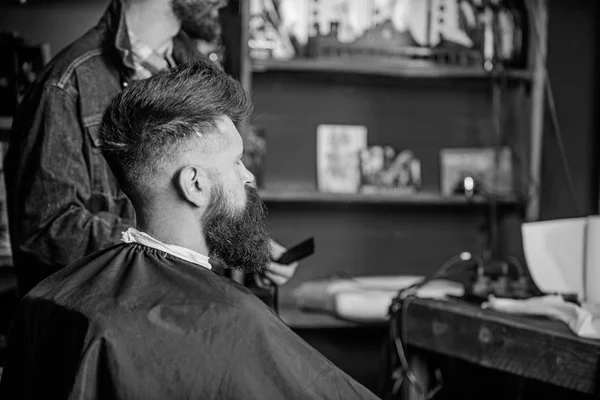 Man with beard covered with black cape sits in hairdressers chair, beauty supplies background. Man with beard client of hipster barbershop. Barbershop concept. Hipster with beard waits for haircut — Stock Photo, Image