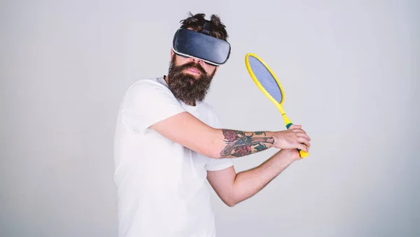 Guy with VR glasses play tennis with racket and ball. Man with beard in VR glasses play tennis, grey background. Hipster on busy face use modern technology for sport games. Virtual sport concept