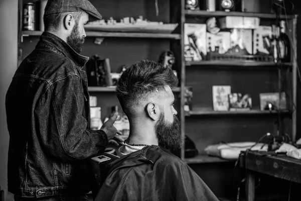 O cliente Hipster tem um novo corte de cabelo. Barbeiro com homem barbudo a olhar para o espelho, fundo da barbearia. Conceito de corte de cabelo. Barbeiro terminou styling. Cliente e mestre profissional verificando resultado — Fotografia de Stock